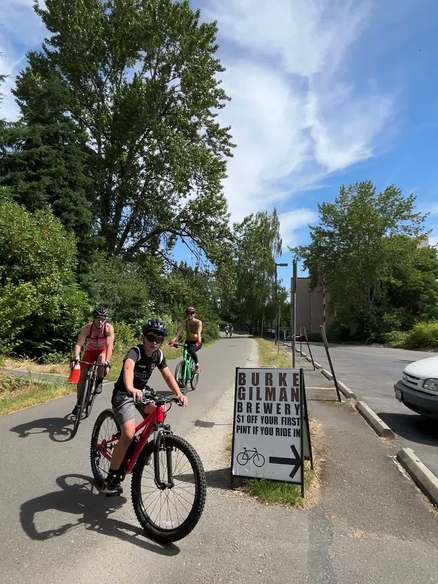 Cyclists riding past the "$1 off your first pint if you ride in" sign on the Burke-Gilman Trail